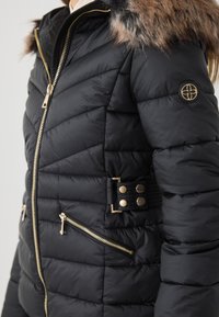 Jachetă puffer neagră cu fermoare aurii, textură matlasată, glugă căptușită cu blană și accente decorative cu nasturi la talie. Două buzunare cu fermoar.