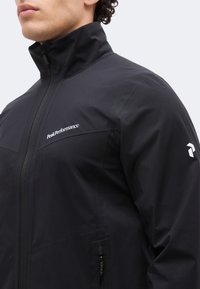 Άτομο που φοράει μαύρο μπουφάν Peak Performance με ψηλό γιακά, φερμουάρ μπροστά και λογότυπο στο στήθος και στο πάνω μέρος του μανικιού.