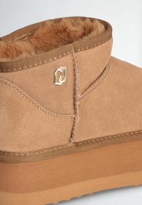 Tan suede ankle boot with a soft fur lining, stitched detailing, and a smooth tan outsole. Features small gold hardware logo accent.