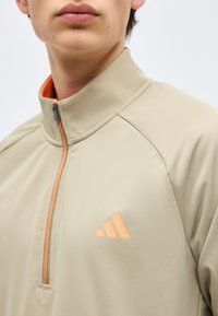 Κοντινό πλάνο ενός ατόμου που φοράει μπεζ μπουφάν με φερμουάρ και πορτοκαλί λογότυπο Adidas στο στήθος καθώς και πορτοκαλί επένδυση στο φερμουάρ.