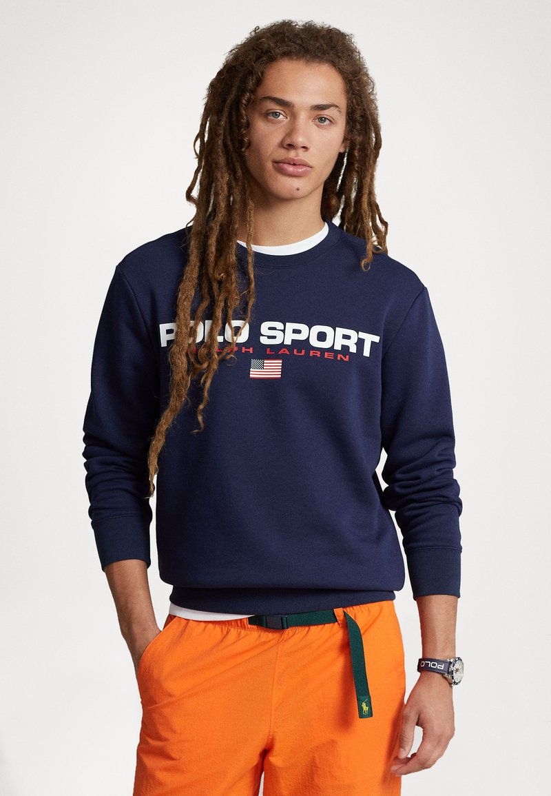 Hanorac bleu marin cu text alb și roșu "POLO SPORT", având un steag american. Asortat cu pantaloni scurți portocalii și o curea verde.