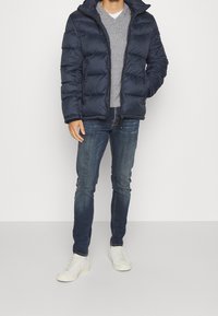 Doudoune navy avec capuche, présentant un design matelassé et une fermeture éclair à l'avant. Portée sur un pull gris à col en V et assortie à un jean noir et des baskets blanches.