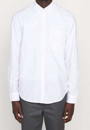 Mand iført en hvid skjorte med lange ærmer og knapper foran med en brystlomme samt mørkegrå bukser, stående foran en ensfarvet baggrund.