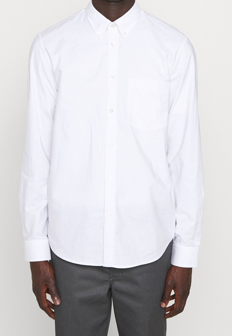 Homme portant une chemise blanche à manches longues avec une poche avant et un pantalon gris foncé, debout devant un fond uni.