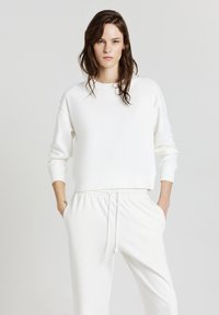 Donna con lunghi capelli castani, vestita con una felpa bianca e pantaloni bianchi con coulisse, in piedi con le mani nelle tasche su uno sfondo neutro.