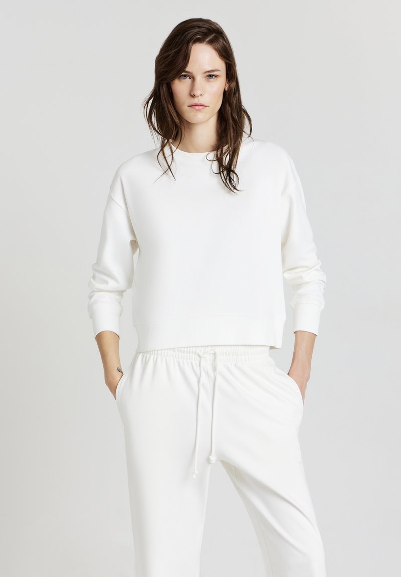 Donna con lunghi capelli castani, vestita con una felpa bianca e pantaloni bianchi con coulisse, in piedi con le mani nelle tasche su uno sfondo neutro.