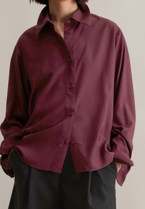 Personne portant une chemise bordeaux ample à manches longues, boutonnée, légèrement rentrée dans un pantalon noir, les mains dans les poches.