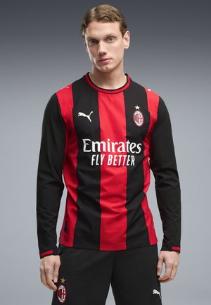 Ung mand iført AC Milan fodboldtrøje med røde og sorte lodrette striber, Emirates Fly Better sponsor og Puma logoer, stående foran en grå baggrund.