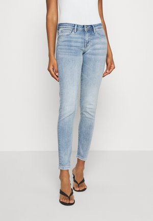 Slim fit lyseblå denim jeans båret med sorte flipflops på en person, der står mod en ensfarvet baggrund.