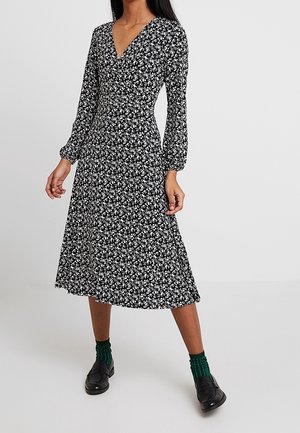 Frau trägt ein schwarzes Midikleid mit weißem Blumenmuster, langen Ärmeln, schwarzen Loafers und grünen Socken, steht vor einem schlichten Hintergrund.