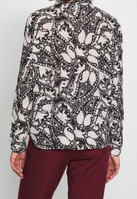 Bluse mit floralem Muster, schwarzem Grund und weißen Designs, langen Ärmeln, kragenartigem Ausschnitt und leichtem, strukturiertem Stoff.