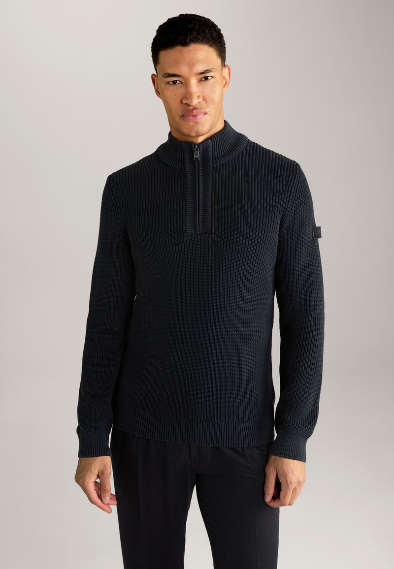 Dunkelblauer, gerippter Strickpullover mit hohem Kragen, halbulloffener Reißverschlussschluss und engen Bündchen, kombiniert mit schwarzen Hosen. Dezente Logodetail am Ärmel.