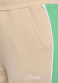 Beige Jogginghosen mit grünem Streifen an der Seite, aus weichem Stoff gefertigt. Verfügt über eine seitliche Tasche und weiße Nähte, mit "s.Oliver" auf der Vorderseite gestickt.