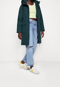 Grøn hætteparka, lysegrøn crop top, slidte lyseblå jeans og hvide sneakers med gule accenter. Casual outfit kombination.
