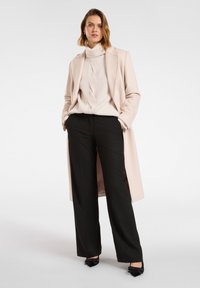 Cappotto lungo rosa pallido su un maglione a coste color crema, abbinato a pantaloni larghi neri e décolleté neri a punta. Design semplice, senza motivi.