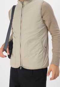 Beige padded vest with a high collar, zip closure, two front pockets, and textured beige sweater sleeves. Features branding on the chest.