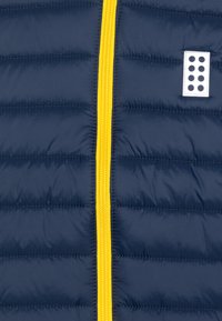 Marineblauer gepolsterter Stoff mit horizontalen Nähten, einem zentralen gelben Reißverschluss und einem kleinen weißen Patch mit schwarzen Punkten auf der rechten Seite.