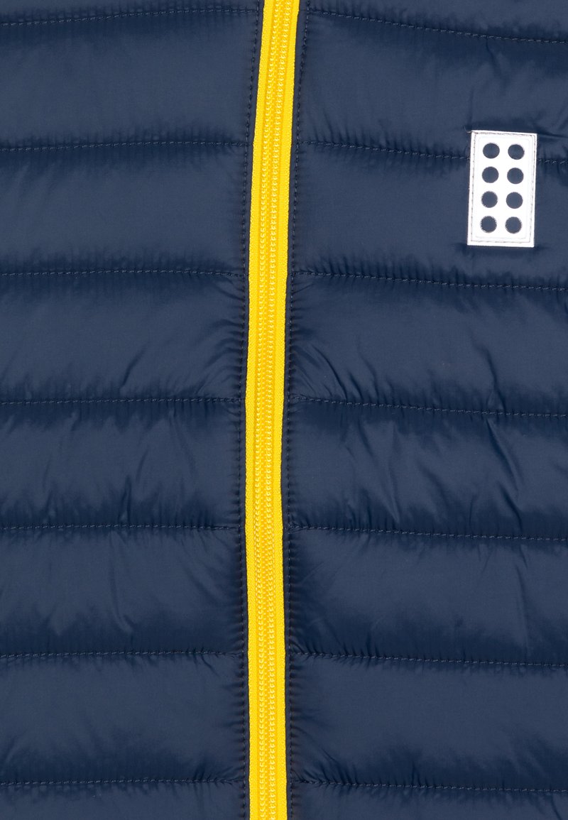 Tessuto imbottito blu navy con cuciture orizzontali, una cerniera centrale gialla e una piccola patch bianca con pois neri sul lato destro.