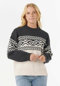 Donna che indossa un maglione lavorato a maglia bianco e nero con motivi a zigzag e a scacchi, abbinato a jeans blu chiaro, in posa con le mani unite.