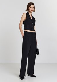 Schwarze, kurze Weste mit Knöpfen und Reverskragen, kombiniert mit hochtaillierten schwarzen Hosen mit einem Gürtel und seitlichen Schlitzen.