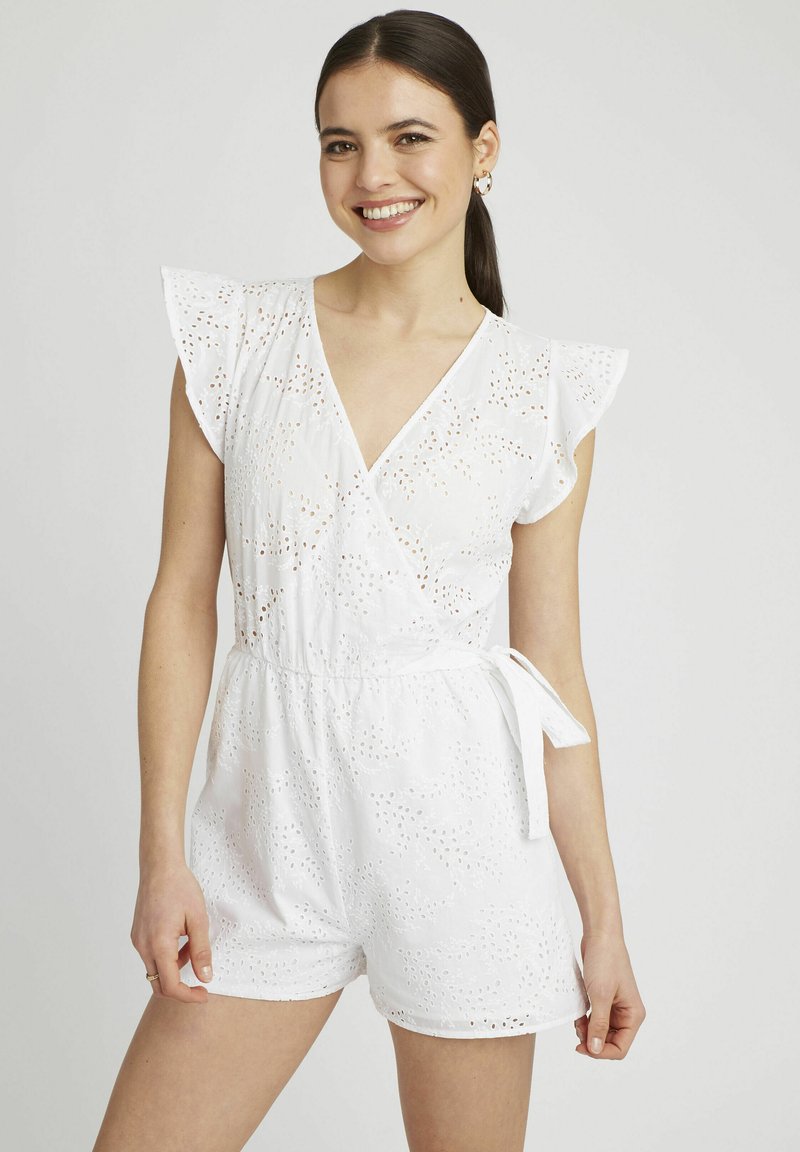 Salopette en coton blanc avec des détails en œillets, décolleté en V, manches flottantes, ceinture à nouer et jambes courtes. Tissu léger et respirant.