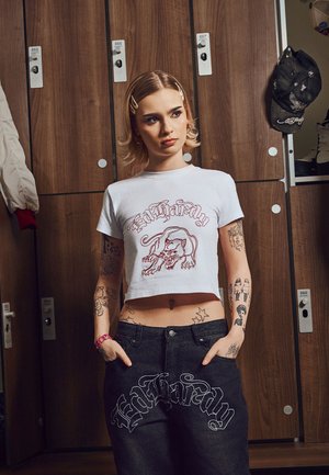 Joven con tatuajes, vestido con camiseta corta blanca y vaqueros oscuros con el logo de "Ed Hardy", de pie frente a taquillas de madera.