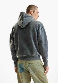 Fiatal férfi hátulnézetben, szürke kapucnis pulóvert visel, amelyen kis hímzett logó található a kapucnin, valamint fakó, két árnyalatú zöld farmernadrágot.