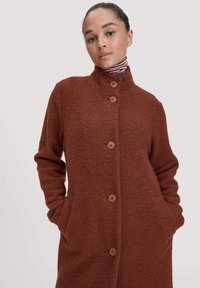 Cappotto di lana marrone con colletto alto, sei bottoni frontali e tasche laterali. Tessuto morbido con una finitura leggermente pelosa.