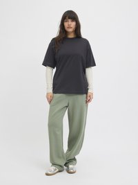 Schwarzes T-Shirt mit kurzen Ärmeln, das über einem hellbeigen Langarmhemd getragen wird, kombiniert mit hellgrünen, lockeren Hosen und silbernen Sneakers.