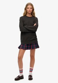 Mujer joven de pie con los brazos cruzados, viste un sudadera negra, una falda de cuadros plisada, calcetas blancas a rayas y mocasines oscuros, todo sobre un fondo blanco.