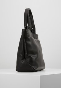 Sac fourre-tout en cuir noir texturé avec double poignées posé sur une surface blanche contre un fond gris uni.