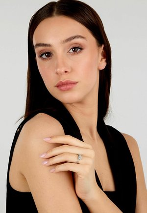 Anello in argento con un design decorativo, il modello indossa un top senza maniche di colore nero, mostrando un incarnato chiaro e smalto per unghie rosa chiaro.