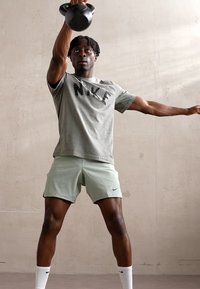 Pelēks Nike T-krekls ar melnu logo, piparmētru zaļas bikses un baltas zeķes. Individuāls, kurš paceļ kettlebellu pret neitrālu sienu fonu.