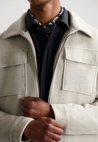 Light gray wool blend jacket with a collar, zip front, and two chest pockets. Worn over a dark shirt with a patterned neckline.