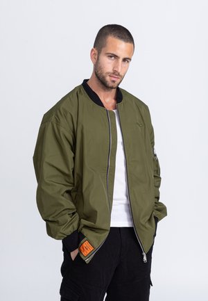 Olijfgroene bomberjack met een zwarte kraag, rits aan de voorkant en een oranje label op de manchet, met een losse pasvorm en een gladde textuur.