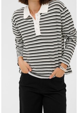 Femme portant un polo à manches longues rayé noir et blanc et un pantalon noir, les mains dans les poches, devant un fond uni.