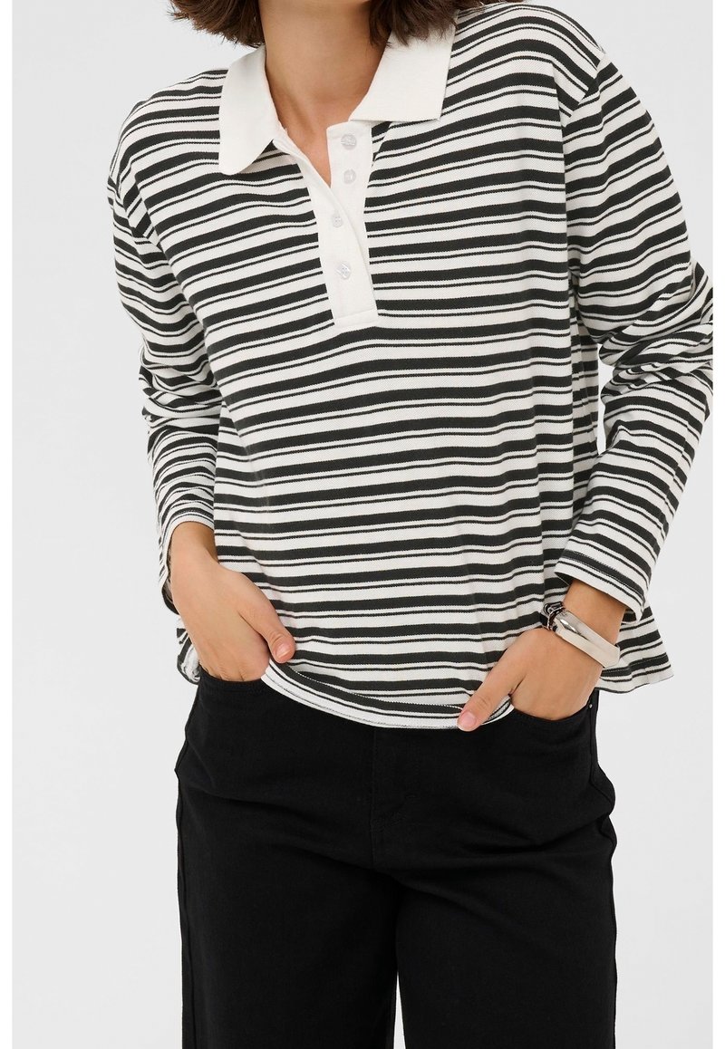 Femme portant un polo à manches longues rayé noir et blanc et un pantalon noir, les mains dans les poches, devant un fond uni.
