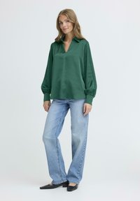 Blusa satinata verde scuro con scollatura a V e maniche a sbuffo, abbinata a jeans azzurri a gamba dritta e ballerine nere a punta.