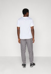 Homme debout de dos, portant une chemise blanche à manches courtes, un pantalon de jogging gris et des chaussures de sport noires, sur un fond clair et uni.
