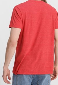 Homme portant un t-shirt rouge uni à manches courtes et un jean bleu clair, vu de dos sur un fond blanc uni.