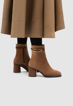 Stivaletti in camoscio marrone con tacco largo, cerniera laterale e fibbia decorativa, indossati con collant neri e gonna marrone abbinata.