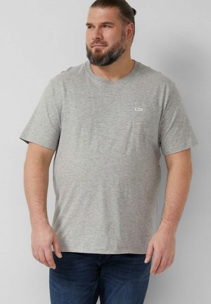 Mann mit Bart und zurückgebundenem Haar, der ein hellgraues kurzärmeliges T-Shirt und dunkelblaue Jeans trägt, vor einem schlichten Hintergrund stehend.