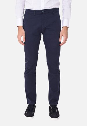Uomo che indossa pantaloni chino blu scuro, camicia bianca con bottoni e sneakers nere con suole bianche, in piedi contro uno sfondo semplice.