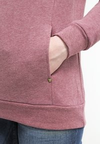 Felpa color rosa con finitura testurizzata, dotata di una tasca laterale e di un piccolo accento metallico a forma di cuore. I polsini sono a coste.