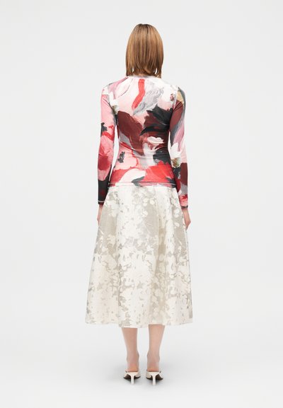 Μπλούζα με μακριά μανίκια και λουλουδένιο μοτίβο σε αποχρώσεις του κόκκινου και ροζ, συνδυασμένη με μακρύ, ανοιχτόχρωμο φούστα με υφασμένο σχέδιο.