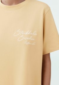 Keltainen puuvillainen t-paita pyöreällä kaula-aukolla, jossa vasemmassa rinnassa on valkoista kalligrafista brodeerausta, jossa lukee "Stockholm Sweden Östermalm".