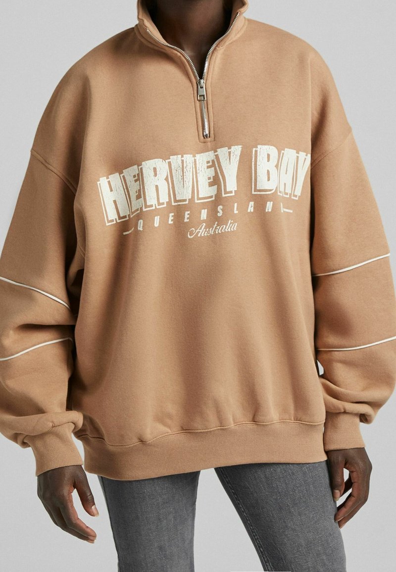 Personne portant un sweat-shirt surdimensionné beige à fermeture éclair quart de zip avec le texte « HERVEY BAY QUEENSLAND Australia », associé à un jean gris.