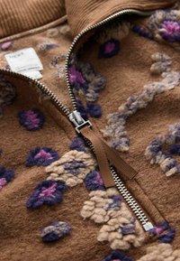 Brązowa polarowa kurtka zapinana na zamek z teksturowanym kwiatowym wzorem w kolorze fioletowym i kremowym, z metalowym zamkiem błyskawicznym i brązowym uchwytem z tkaniny.