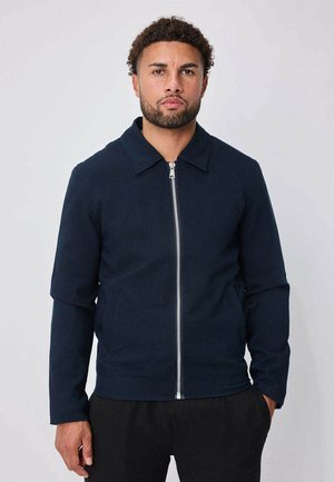 Homme aux cheveux courts et bouclés avec une barbe, portant une veste bleu marine zippée et un pantalon noir, debout devant un fond uni.