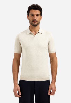 Man met kort krullend haar, gekleed in een beige polo met korte mouwen en een zwarte broek, staand tegen een witte achtergrond.